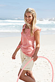 A young woman on a beach with a hula-hoop wearing a pink top and shorts