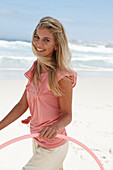 A young woman on a beach with a hula-hoop wearing a pink top and shorts