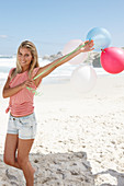 A young woman on a beach with balloons wearing a pink top and denim shorts