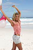 A young woman on a beach with balloons wearing a pink top and denim shorts