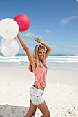 A young woman on a beach with balloons wearing a pink top and denim shorts