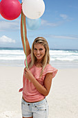 A young woman on a beach with balloons wearing a pink top and denim shorts