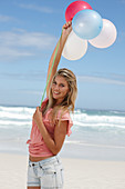 A young woman on a beach with balloons wearing a pink top and denim shorts