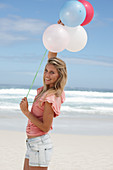 A young woman on a beach with balloons wearing a pink top and denim shorts