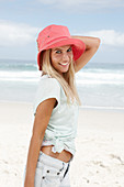 A blonde woman on the beach wearing a light t-shirt and a red hat