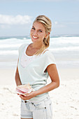 A blonde woman on the beach wearing a light t-shirt and holding a shell