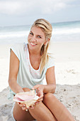 A blonde woman on the beach wearing a light t-shirt and holding a shell