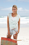 A blonde woman on the beach wearing a light t-shirt and denim shorts holding a wicker bag