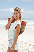 A blonde woman on the beach wearing a light t-shirt and denim shorts holding a wicker bag