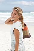 A blonde woman on the beach wearing a light t-shirt and holding a wicker bag