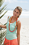 A blonde woman on a beach wearing a scarf, a turquoise top and a salmon pink skirt
