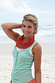 A blonde woman on a beach wearing a turquoise top and a red necklace