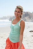 A blonde woman on a beach wearing a turquoise top and a salmon-pink skirt