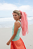 A blonde woman on a beach with a scarf wearing a turquoise top and a salmon-pink skirt