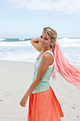 A blonde woman on a beach with a scarf wearing a turquoise top and a salmon-pink skirt