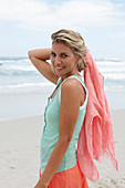 A blonde woman on a beach with a scarf wearing a turquoise top and a salmon-pink skirt