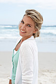 A blonde woman on a beach wearing a turquoise top and a white cardigan