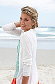 A blonde woman on a beach wearing a turquoise top and a white cardigan