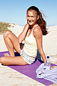 A brunette woman sitting on a beach mat wearing a top and shorts