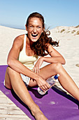 A brunette woman sitting on a beach mat wearing a top and shorts