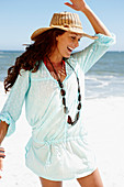 A brunette woman wearing a hat, a necklace and a light-blue beach dress by the sea