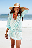 A brunette woman wearing a hat and a light-blue beach dress by the sea