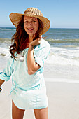 A brunette woman wearing a hat and a light-blue beach dress by the sea