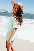 A brunette woman wearing a hat and a light-blue beach dress by the sea