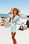 A brunette woman wearing a hat and a light-blue beach dress by the sea