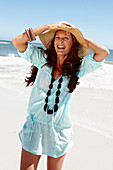 A brunette woman wearing a hat, a necklace and a light-blue beach dress by the sea