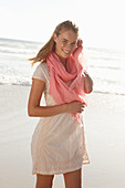 A blonde woman by the sea wearing a salmon-pink scarf and a white dress