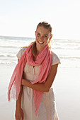 A blonde woman by the sea wearing a salmon-pink scarf and a white dress