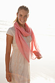 A blonde woman by the sea wearing a salmon-pink scarf and a white dress