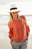 Junge blonde Frau mit Hut in orangefarbener Bluse und heller Hose am Meer