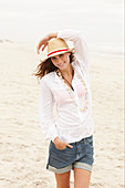 A brunette woman wearing a hat, a white blouse and denim shorts