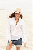 A brunette woman wearing a hat, a white blouse and denim shorts