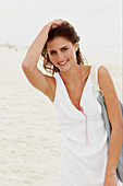 A brunette woman by the sea with a bag wearing a white summer dress