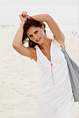 A brunette woman by the sea with a bag wearing a white summer dress