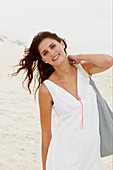 A brunette woman by the sea with a bag wearing a white summer dress