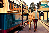 A young couple next to a tram, her wearing a summer dress and him wearing a jacket and a jumper