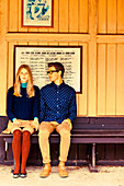 Junges Paar in Rock, Strümpfen, Shirt und Hose sitz händchenhaltend auf einer Bank
