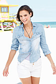 A young woman on a beach wearing a denim shirt and white shorts