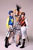 A blonde woman wearing a printed blouse, a matching skirt and a hat with two men in jockey's outfits