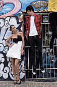 Junge Frau in schwarz-weißem Partykleid und junger Mann in kariertem Hemd und Jeans vor Wand mit Graffiti