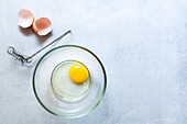 Aufgeschlagenes Ei in Glasschale, daneben Schneebesen und Eierschalen