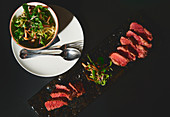 Slices of tasty meat and bowl with arugula and mushroom salad on black background