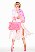 A blonde woman wearing a pink blouse and a fringed skirt with a bag holding a pink dog