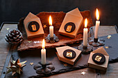 Adventskranz mit nummerierten Papierhäuschen auf Baumrinde und Schieferplatte mit brennenden Kerzen