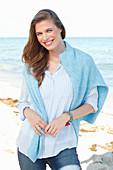 A young woman by the sea wearing a light blue shirt, jumper and jeans