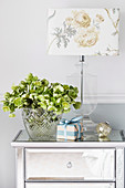 Christmas roses in a glass vase next to a table lamp with a floral motif on a silver bedside table
